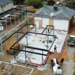 Aerial view of a construction site showing steel frame erection on a concrete slab, with workers and equipment visible.