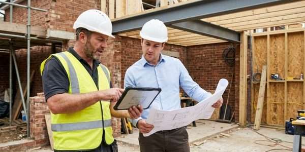 Two construction workers in hard hats reviewing building plans and a tablet on a construction site